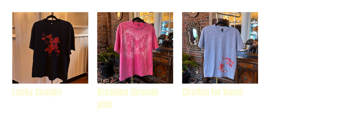 Three graphic t-shirts displayed side-by-side on hangers in boutique settings, labeled 'Lucky Cowgirl' in black with red graphic, 'Breaking through pink' in bright pink with butterfly graphic, and 'Circling for home' in light blue with red graphic.