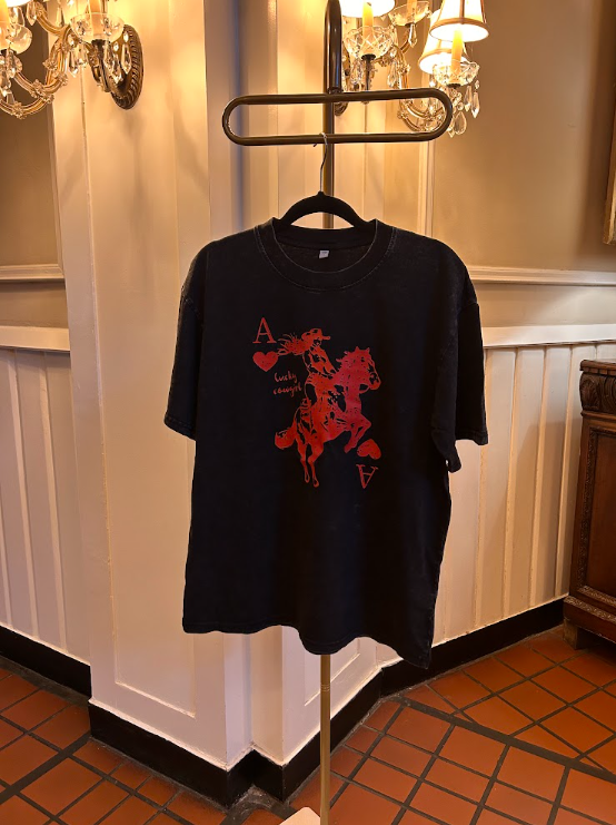 A black oversized graphic T-shirt hanging on a black metal stand in a vintage room with white wainscoting and terracotta tile floors. The shirt features a red graphic of an Ace of Hearts playing card with a cowboy riding a horse.